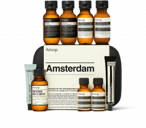 A travel amenities kit, with a black bag featured in the background. The bag is labled "Aesop, Amsterdam". Seven small bottles in the foreground containing toiletries including shampoo and conditioner. There are also two tubes, including a tube of toothpaste.