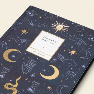 A dark blue journal with a cosmic pattern featuring eyes, hands, stars, moons, and hourglasses in a lighter shade of blue. Two gold stars, two gold crescent moons, one sun, and one snake are also featured on the design. A white box in the center reads 'Jennifer Morales, mid-year diary'.