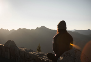 A silhouette of a person sitting on a cliff face, looking at mountains