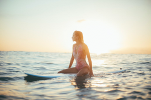 A woman in a pink one-piece bathing suit sits on a blue surfboard in the middle of the ocean with the sun behind her.