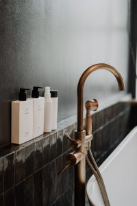 Brass bath faucet with cream bottles of toiletries next to it in a dark gray bathroom